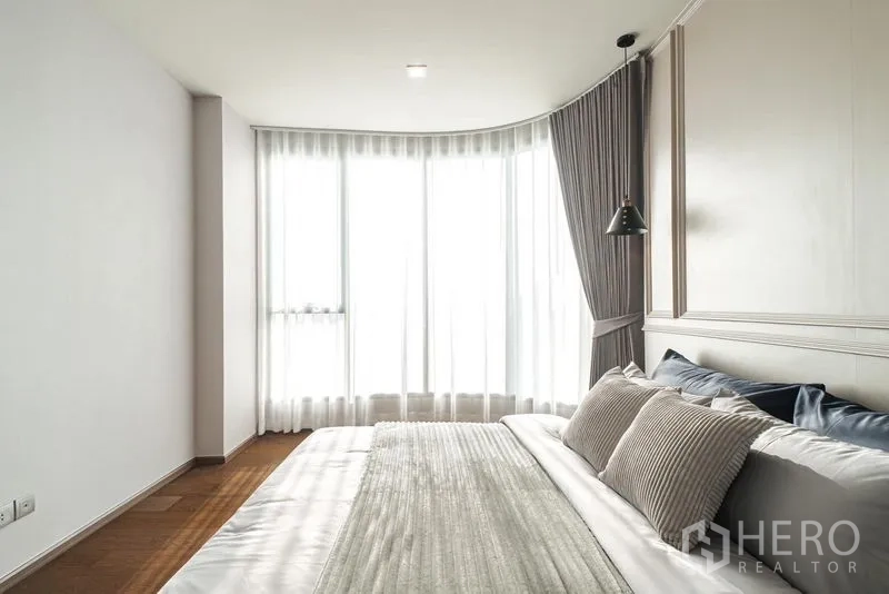 Condominium for rent in Khlong Toei, Bangkok - Sunlit bedroom with a king bed, sheer curtains, and a curved floor-to-ceiling window.
