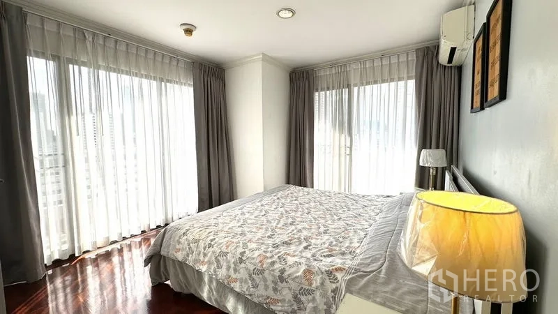 Condominium for rent or sale in Watthana, Bangkok - Corner primary bedroom with large wraparound windows, sheer curtains, and bedside lamp.