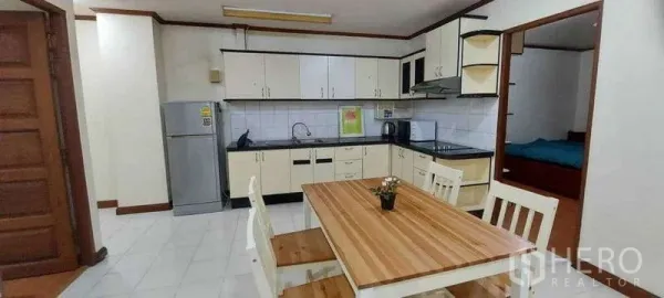 Condominium for rent in Khlong Toei, Bangkok - Open kitchen with white cabinets, tiled backsplash and a wooden dining table beside a doorway to a bedroom.