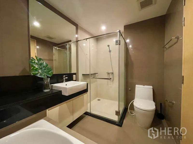 Condominium for rent or sale in Khlong Toei, Bangkok - Second bathroom with glass-enclosed shower, toilet, and wide vanity mirror.