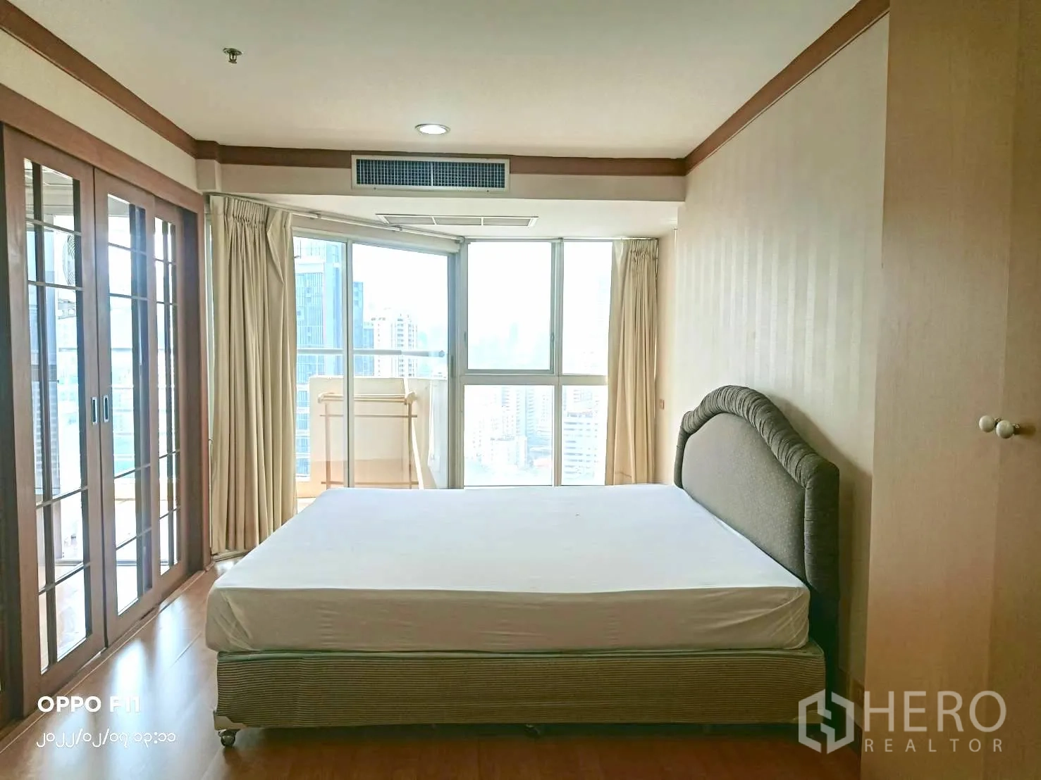 Condominium for rent in Khlong Toei, Bangkok - Sunlit bedroom with large windows, city views, beige curtains and a queen bed.