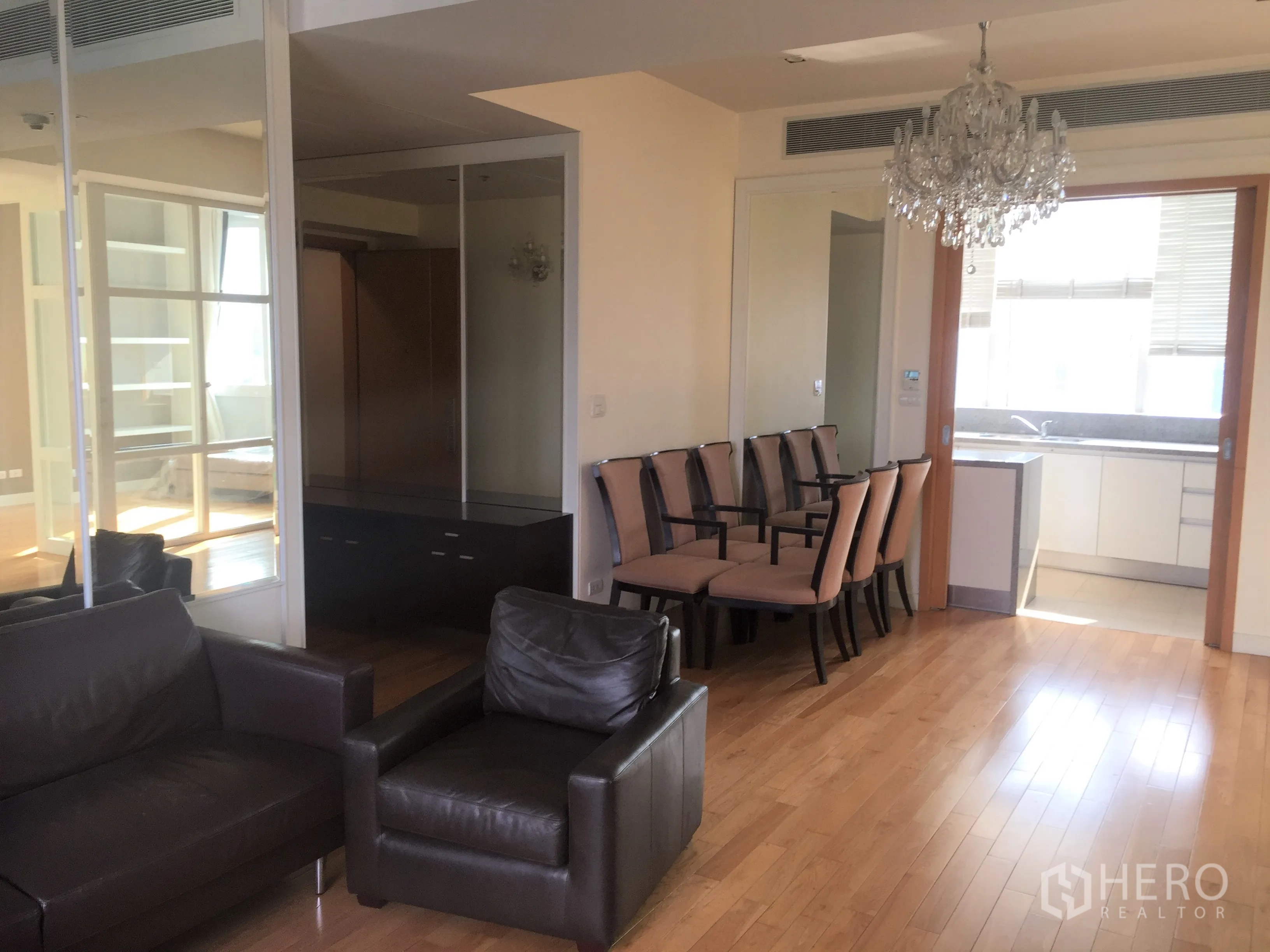 Condominium for rent in Khlong Toei, Bangkok - Open-plan living and dining with crystal chandelier, mirrored wall, and sliding door to the kitchen.