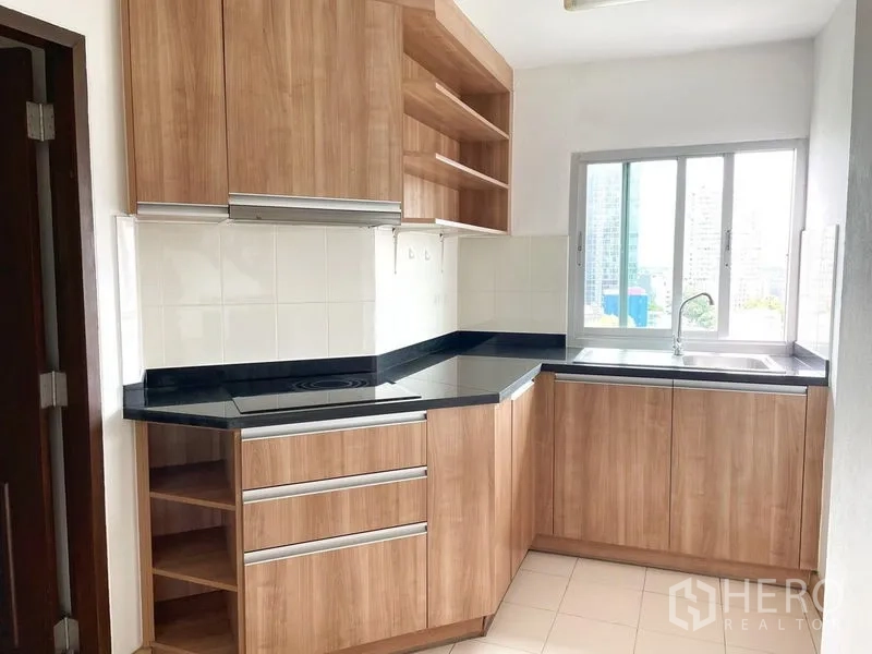 Condominium for rent in Khlong Toei, Bangkok - Corner kitchen with wood cabinets, black stone counters, electric hob and window over the sink.