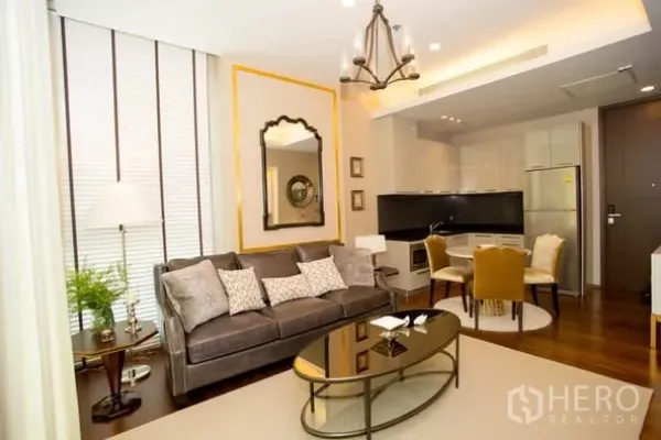 公寓 出租在Watthana, Bangkok - 通罗公寓开放式客厅，灰色沙发、金色饰边镜与吊灯，连接现代厨房与餐区。