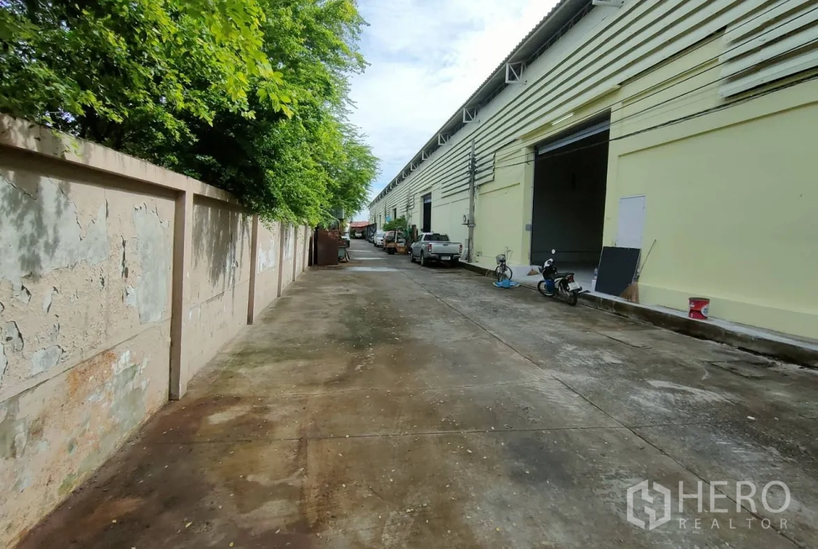 货栈 出租在Bang Sao Thong, Samut Prakan - 仓库排旁的通道，装卸门敞开，路旁树木成荫
