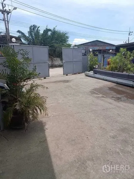 Warehouse for sale in Samut Sakhon - Wide concrete yard inside the compound with sliding metal gates and potted plants along the boundary.