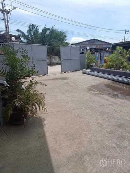 Warehouse for sale in Samut Sakhon - Wide concrete yard inside the compound with sliding metal gates and potted plants along the boundary.