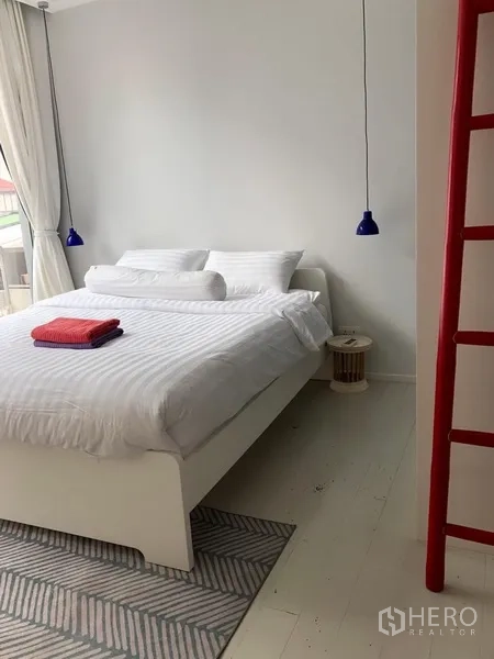Condominium for rent or sale in Watthana, Bangkok - Bright master bedroom with white bed, blue pendant lights, red ladder shelf and floor-to-ceiling curtains.