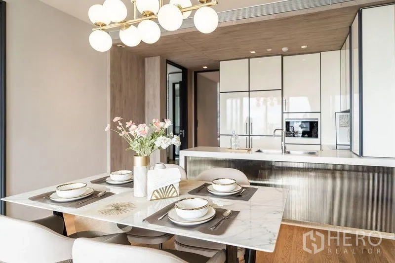 Condominium for rent in Khlong Toei, Bangkok - Dining table set for four with modern chandelier and kitchen island in the background.