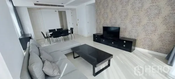 Condominium for rent in Pathum Wan, Bangkok - Living room with L-shaped gray sofa, TV console, feature wall, black coffee table and adjacent dining space.