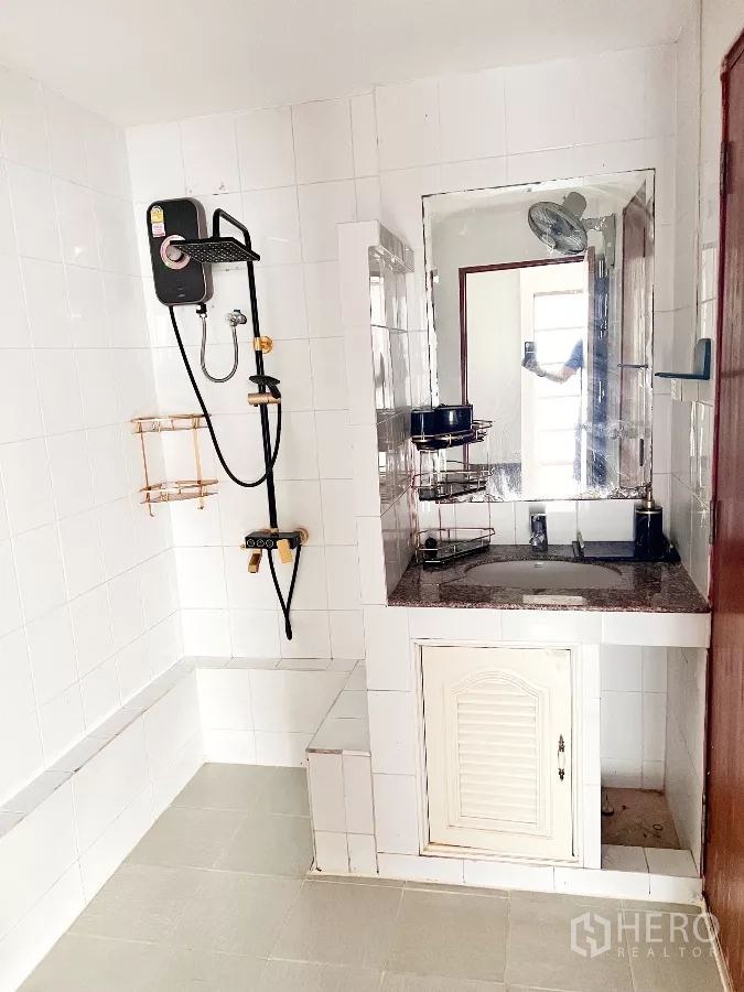 Townhouse for rent or sale in Phra Khanong, Bangkok - White-tiled bathroom with electric shower, vanity and large mirror.