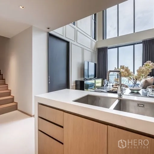 Condominium for rent in Khlong Toei, Bangkok - Open kitchen with island sink and wood cabinetry looking toward the living room and expansive glass facade, with stairs nearby.