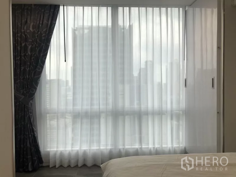 คอนโด ให้เช่าในSathon, Bangkok - มุมห้องนอนพร้อมหน้าต่างสูงจรดเพดาน ผ้าม่านโปร่ง และวิวสกายไลน์บางส่วน