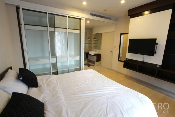 Condominium for rent or sale in Watthana, Bangkok - Primary bedroom with glass-front built-in wardrobe, wall-mounted TV and access to an ensuite vanity in a Thonglor condominium.