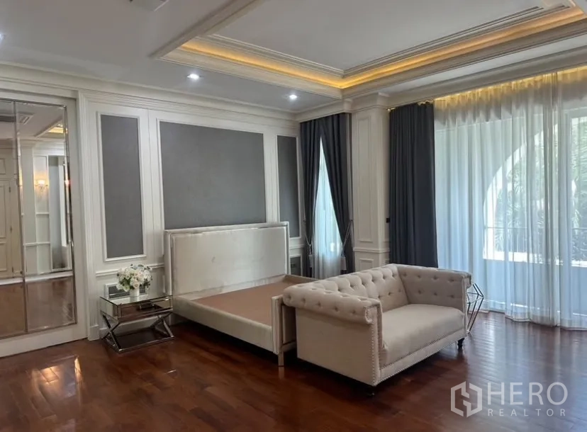 Detached House for rent or sale in Prawet, Bangkok - Spacious master bedroom with wood flooring, paneled walls, bedframe, tufted sofa, and mirrored wardrobe.