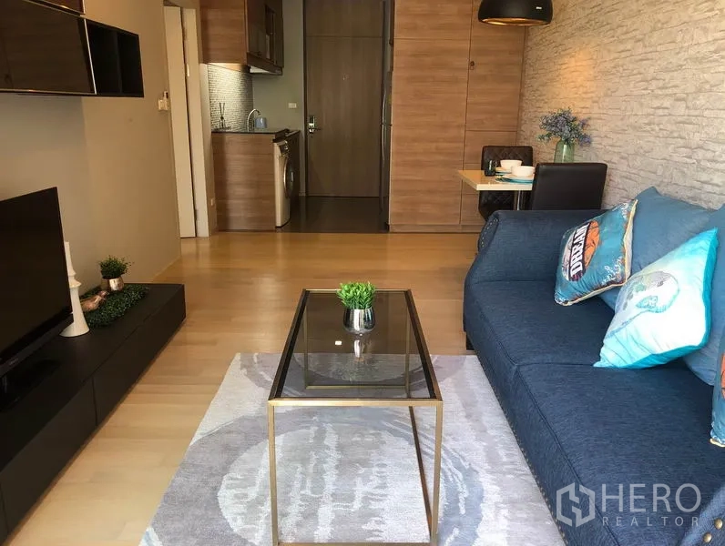 Condominium for rent in Bangkok - Modern living room with blue sofa, glass coffee table, TV console, and open kitchenette with washer in a Bangkok condo.