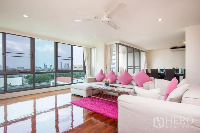 Condominium for rent in Bang Rak, Bangkok - Bright corner living room with floor-to-ceiling windows, skyline and park views, white sofas and pink cushions.