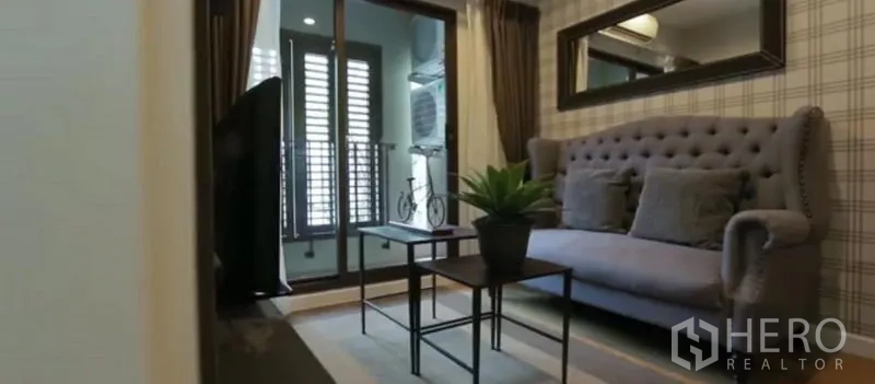 Condominium for rent in Khlong Toei, Bangkok - Living room featuring a tufted gray sofa, coffee table, large mirror, and balcony access.