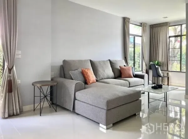 Detached House for rent in Prawet, Bangkok - Corner living room with grey sectional sofa, large windows, curtains and garden views.