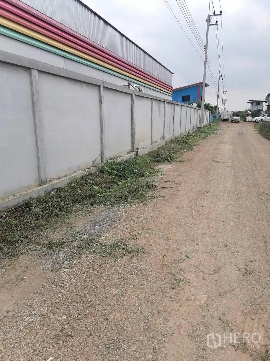 仓库 for sale in Lam Luk Ka, Pathum Thani - 仓库旁的碎石道路与混凝土围墙、电线杆沿线延伸。