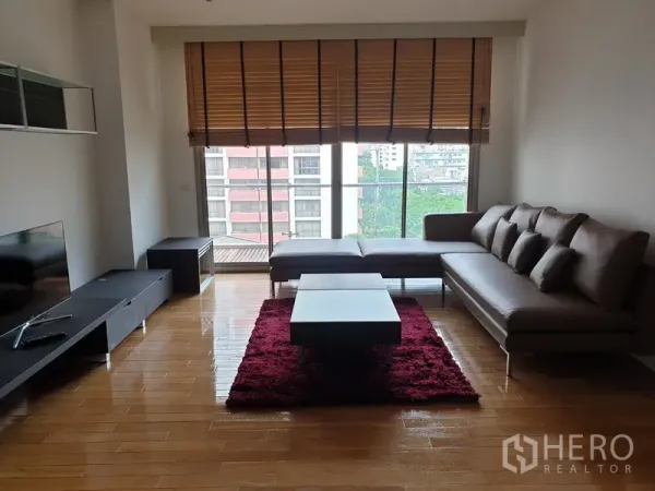 公寓 出租在Bang Rak, Bangkok - 客厅设L形沙发、电视柜与红色地毯茶几，落地窗望城市景观与阳台。