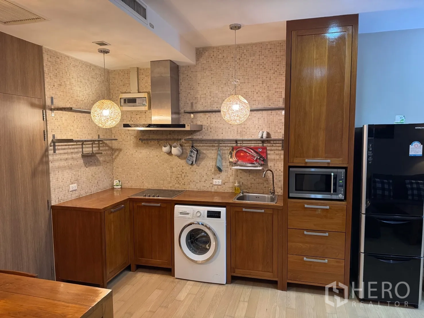 公寓 for rent or sale in Watthana, Bangkok - 开放式厨房具木质橱柜、电灶、烟机、水槽、微波炉、冰箱与室内洗衣机，吊灯点缀其间。