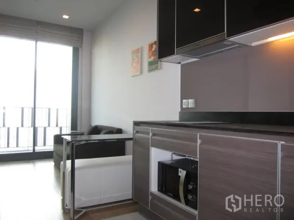Condominium for rent in Khlong Toei, Bangkok - Modern kitchenette with built-in microwave and cabinets beside a sofa and large balcony doors providing natural light.