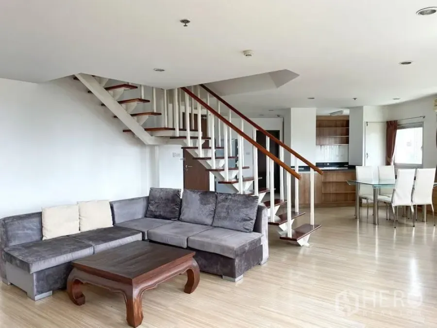 Condominium for rent in Khlong Toei, Bangkok - Bright duplex living area with gray sectional sofa, wood floors, open staircase and dining space beside the kitchen.