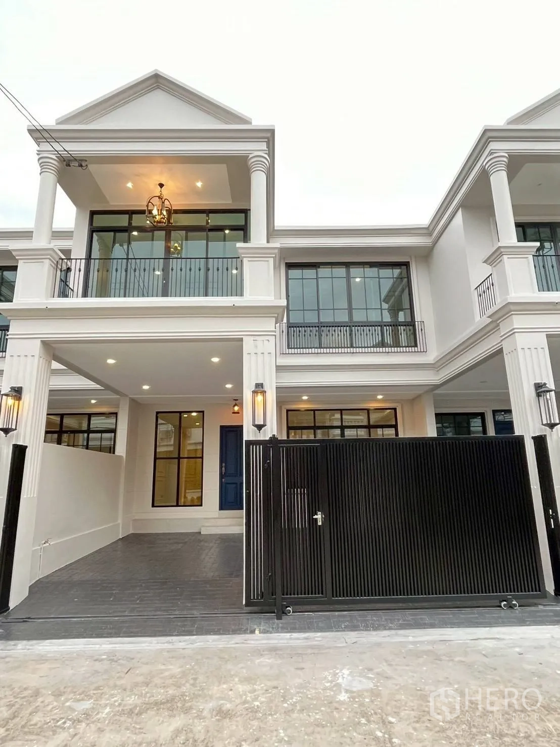 ทาวน์เฮาส์ for rent in Khlong Toei, Bangkok - ด้านหน้าทาวน์เฮ้าส์สีขาวสไตล์คลาสสิก มีระเบียง ไฟสว่าง และรั้วเลื่อนสีดำ.