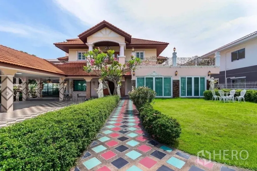 Detached House for rent or sale in Bang Khen, Bangkok - Colorful tiled walkway leading to a two‑storey detached house with red tile roof, balcony, statues, and a large manicured lawn.