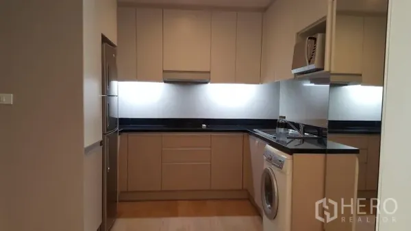 Condominium for rent in Khlong Toei, Bangkok - U-shaped kitchen with beige cabinetry, black stone countertop, fridge and washing machine.