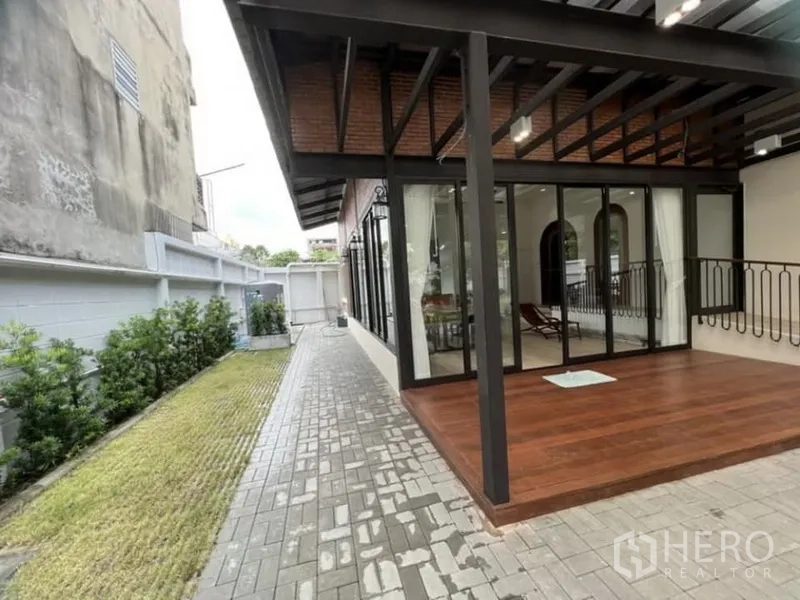 独立式住宅 出租在Ratchathewi, Bangkok - 侧院铺砖步道，金属顶棚下的木露台与玻璃客厅。