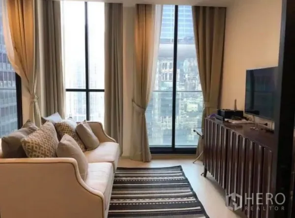 Condominium for rent in Pathum Wan, Bangkok - Bright living room with large windows and balcony door, sofa and media console overlooking the skyline.