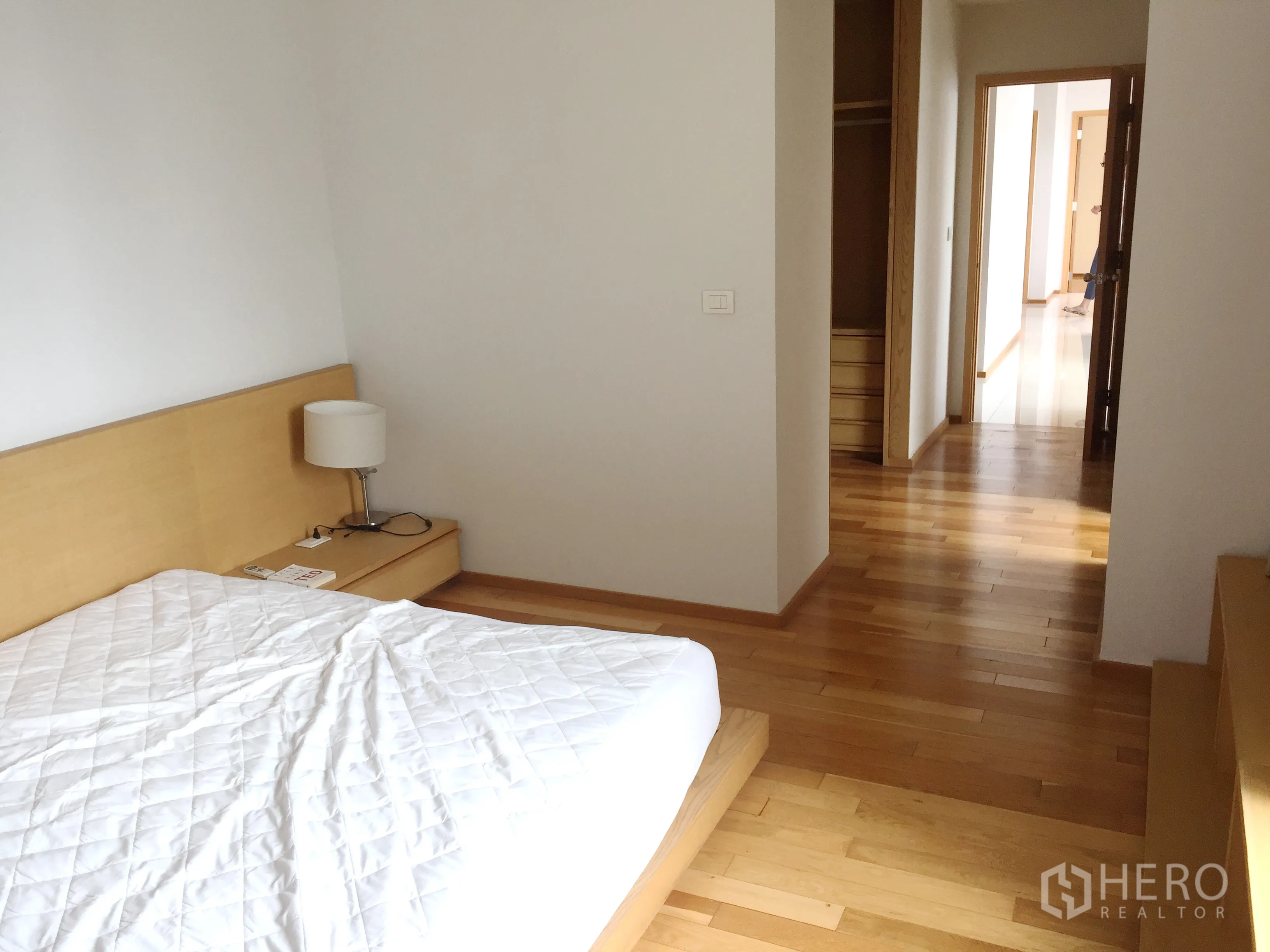 Condominium for rent in Khlong Toei, Bangkok - Minimalist bedroom with platform bed, bedside table, and corridor to built-in wardrobe on hardwood floors.