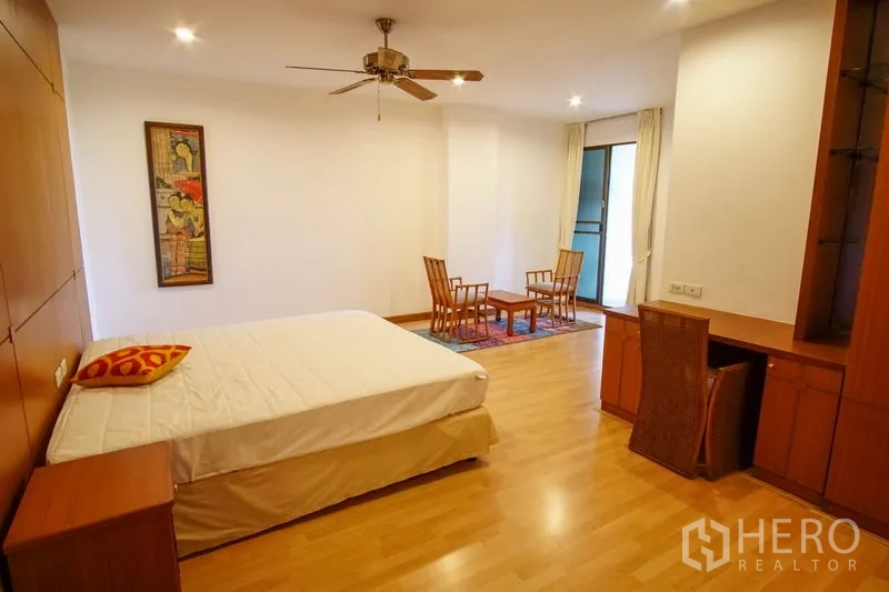 Apartment for rent in Watthana, Bangkok - Oversized bedroom with seating area, wood floors, ceiling fan, built-in desk, and balcony access.