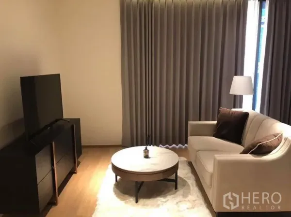 Condominium for rent in Khlong Toei, Bangkok - Living room with TV console, round coffee table, and sofa facing full-length curtains.