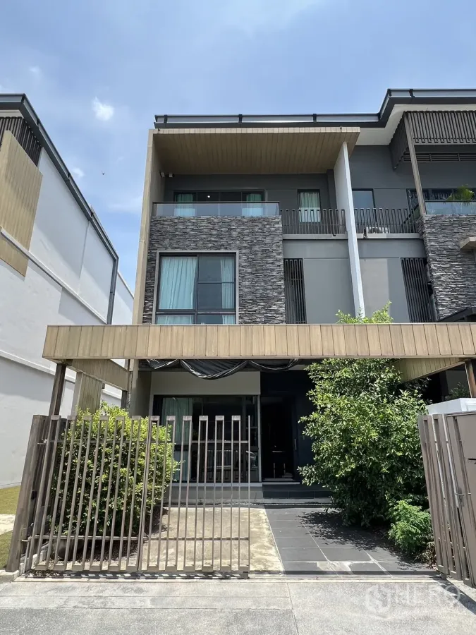 Townhouse for rent in Lat Phrao, Bangkok - Modern three-storey townhouse facade with stone cladding, balcony and gated driveway with greenery.