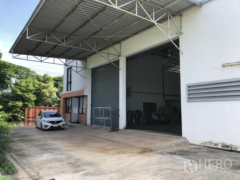 Factory for rent or sale in Lat Lum Kaeo, Pathum Thani - Covered loading bay with extra-wide roller shutter and parking apron beside greenery.