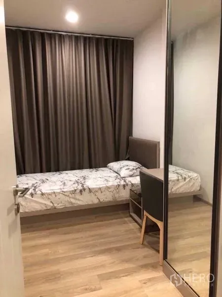 Condominium for rent in Khlong Toei, Bangkok - Compact second bedroom with single bed, desk chair, mirrored wardrobe, blackout curtains, and wood flooring.