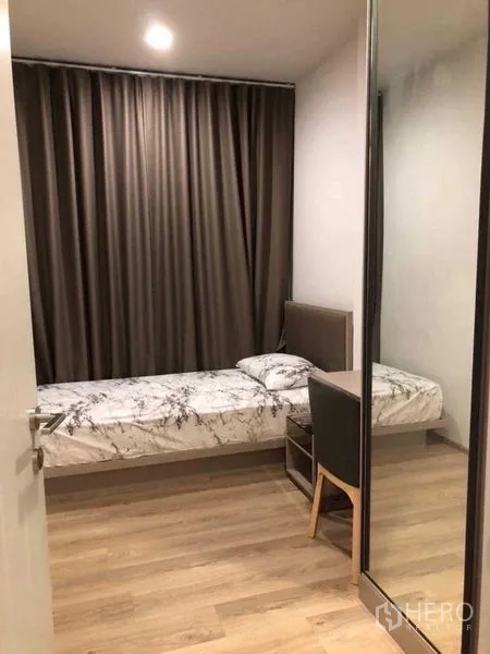 Condominium for rent in Khlong Toei, Bangkok - Compact second bedroom with single bed, desk chair, mirrored wardrobe, blackout curtains, and wood flooring.