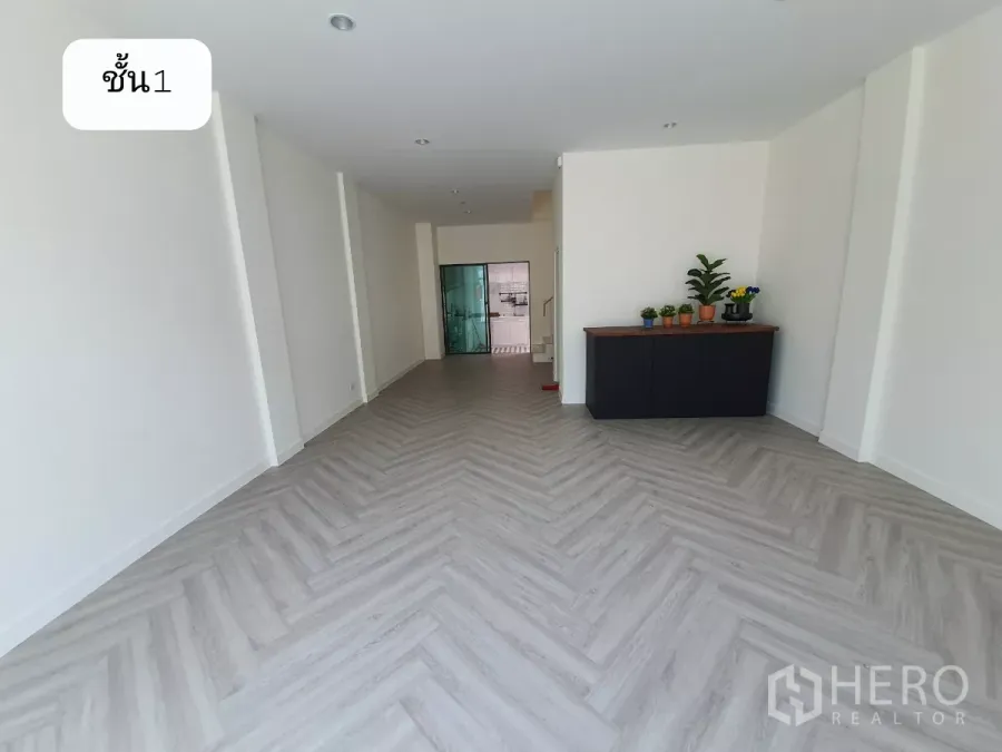 带办公室的家 出租或出售在Prawet, Bangkok - 一层开放式空间，鱼骨拼木纹地板、白墙，侧边矮柜与绿植，后方玻璃隔断。