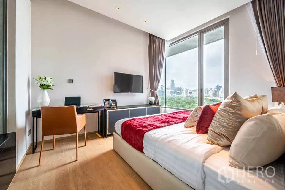 公寓 出租或出售在Bang Rak, Bangkok - 卧室带书桌与墙挂电视，落地窗俯瞰绿意与曼谷天际线。