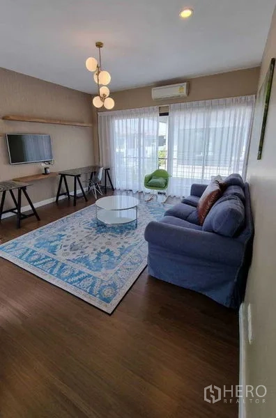 Detached House for rent or sale in Prawet, Bangkok - Living room with blue sofa, TV, desk and large windows with sheer curtains.