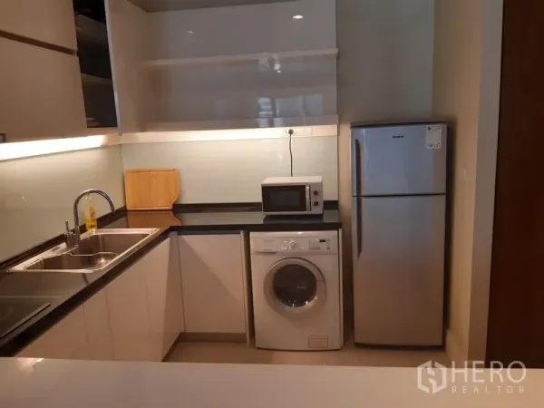 Condominium for rent in Khlong Toei, Bangkok - Compact L-shaped kitchen featuring double sink, black countertop, washing machine, microwave and fridge.