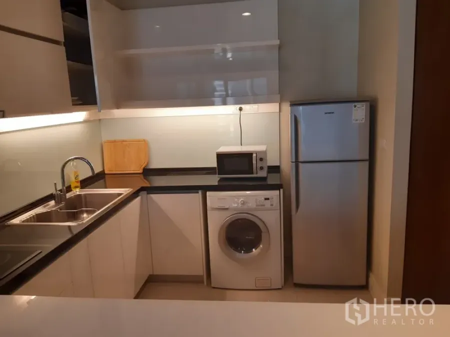 Condominium for rent in Khlong Toei, Bangkok - Compact L-shaped kitchen featuring double sink, black countertop, washing machine, microwave and fridge.