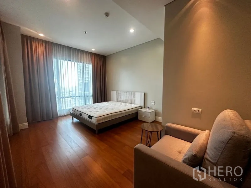 Condominium for rent in Khlong Toei, Bangkok - Spacious bedroom with large window, bed, armchair and warm wooden flooring.
