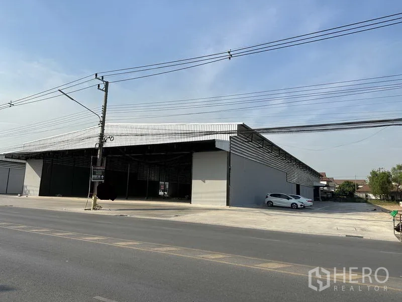 Warehouse for rent in Lam Luk Ka, Pathum Thani - Street-facing view of a large metal-clad warehouse with wide open bay and broad concrete forecourt.