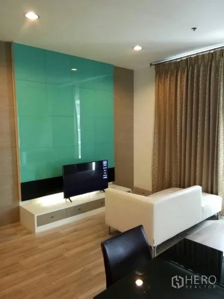 公寓 出租在Ratchathewi, Bangkok - 明亮客厅，白色沙发、电视柜与青绿色玻璃背景墙，木地板与落地窗帘。