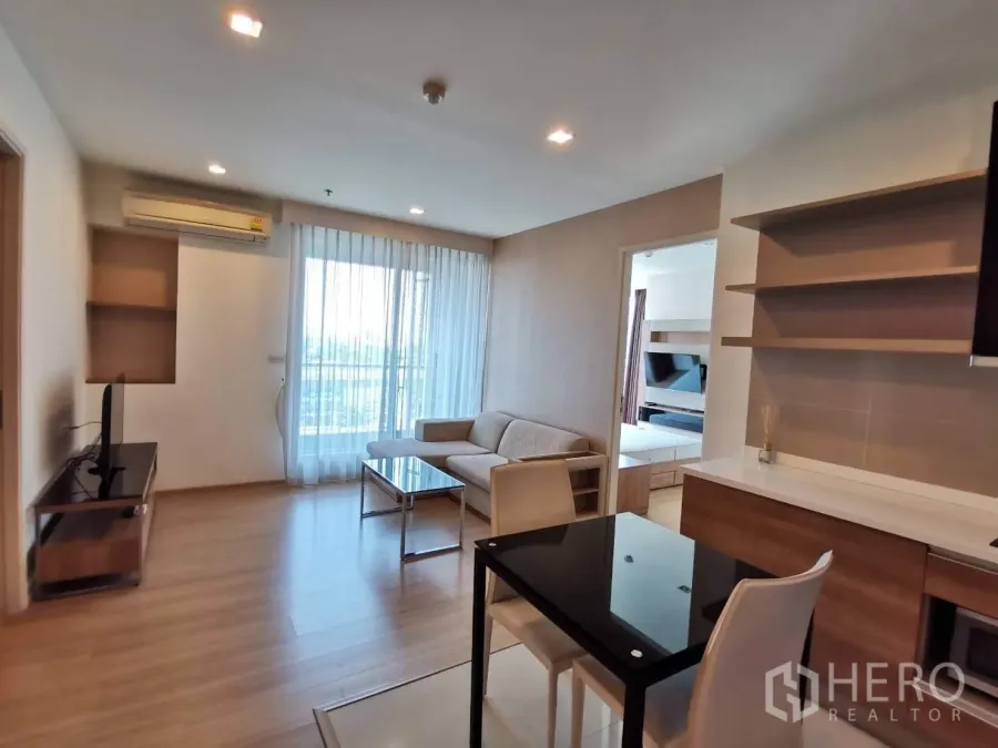 Condominium for rent or sale in Khlong Toei, Bangkok - Dining and lounge area with TV stand, full-height window to balcony, and open shelving.