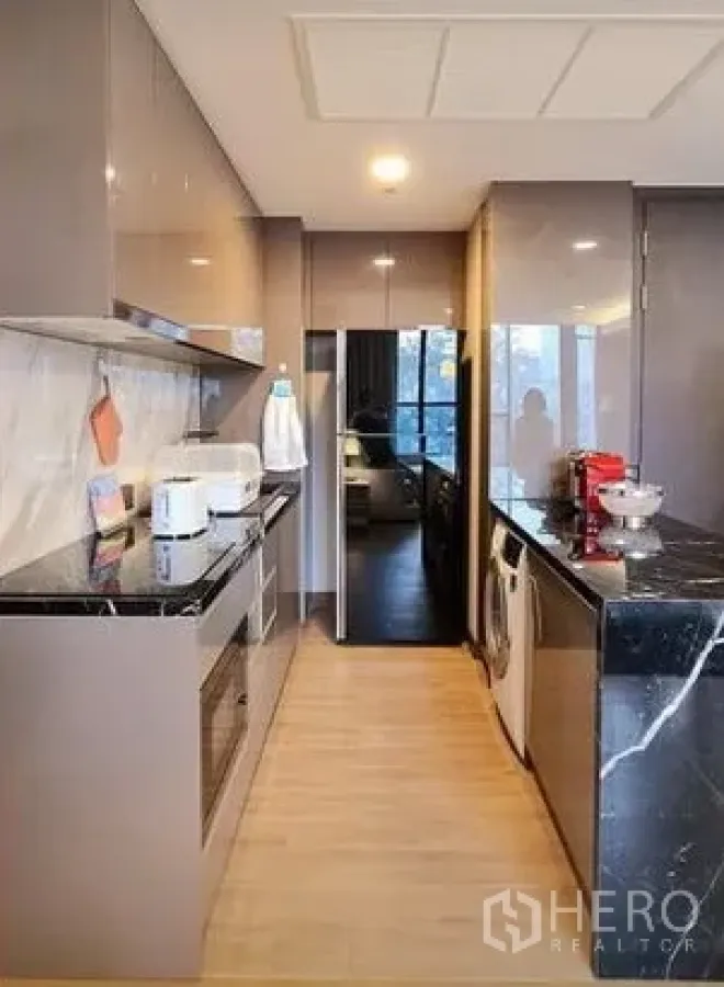 Condominium for rent in Khlong Toei, Bangkok - Modern galley kitchen with glossy cabinets, black stone countertop, built‑in oven, fridge and washer.